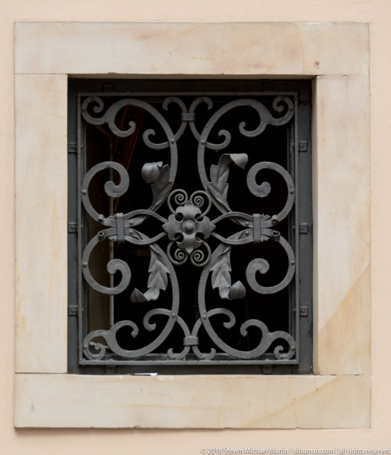 Black wrought-iron window grille with curling vines, leaves, and central floral rosette in a pale stone frame.