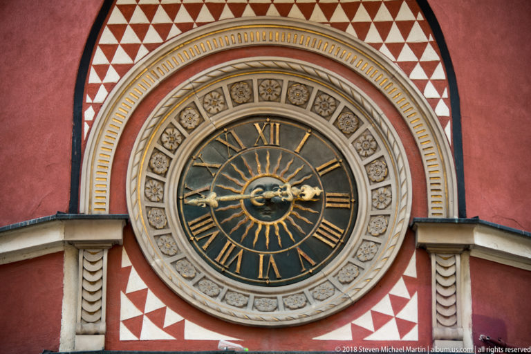 Decorative clock with Roman numerals, gilded hands, and sunburst center set in a red-and-white patterned facade.