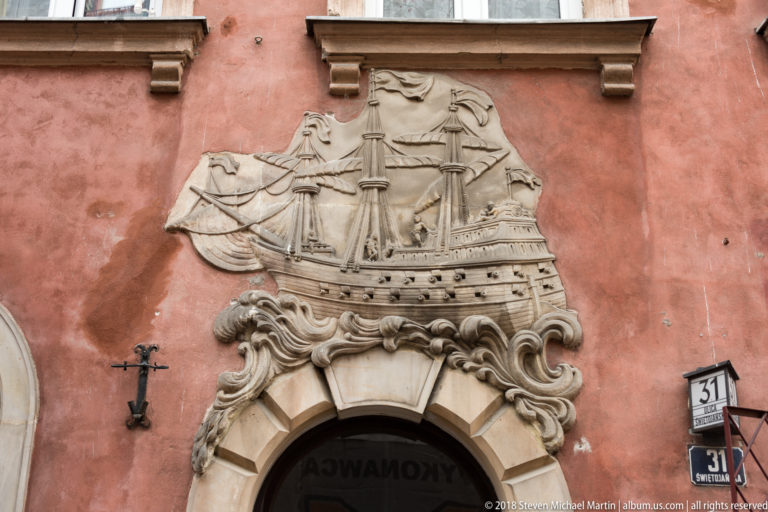 Stone relief of a multi-masted sailing ship above an arched doorway on a pink Old Town facade.