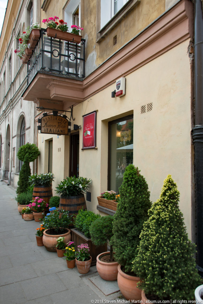 Historic Warsaw storefront with restaurant entrance surrounded by numerous potted shrubs, flowers, and clipped greenery.