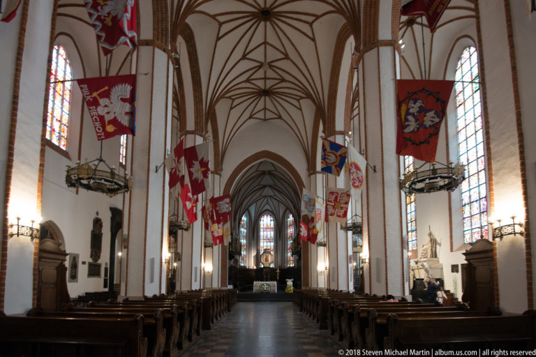 St. John's Archcathedral nave with brick piers, Gothic rib vaults, hanging banners, wooden pews, and sanctuary beyond.