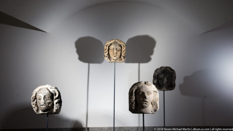 Four carved stone heads on thin stands cast large shadows on a wall in a cathedral crypt display.