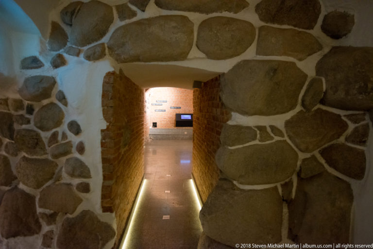 Low stone archway leading into a narrow brick-lined crypt passage illuminated by floor-level lights.