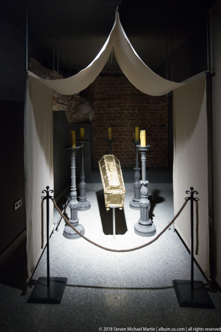 Spotlighted crypt display with an open book or inscribed object between candle stands beneath a white canopy.
