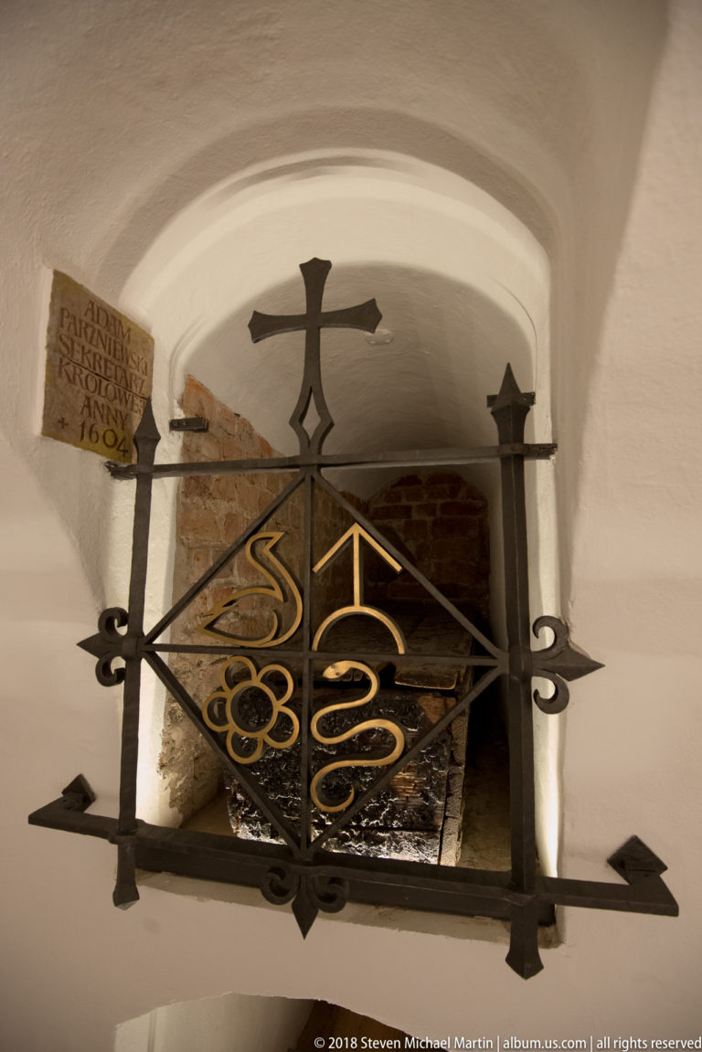 Iron-gated arched memorial niche containing a stone fragment and inscription, with decorative crosses and gilded motifs.