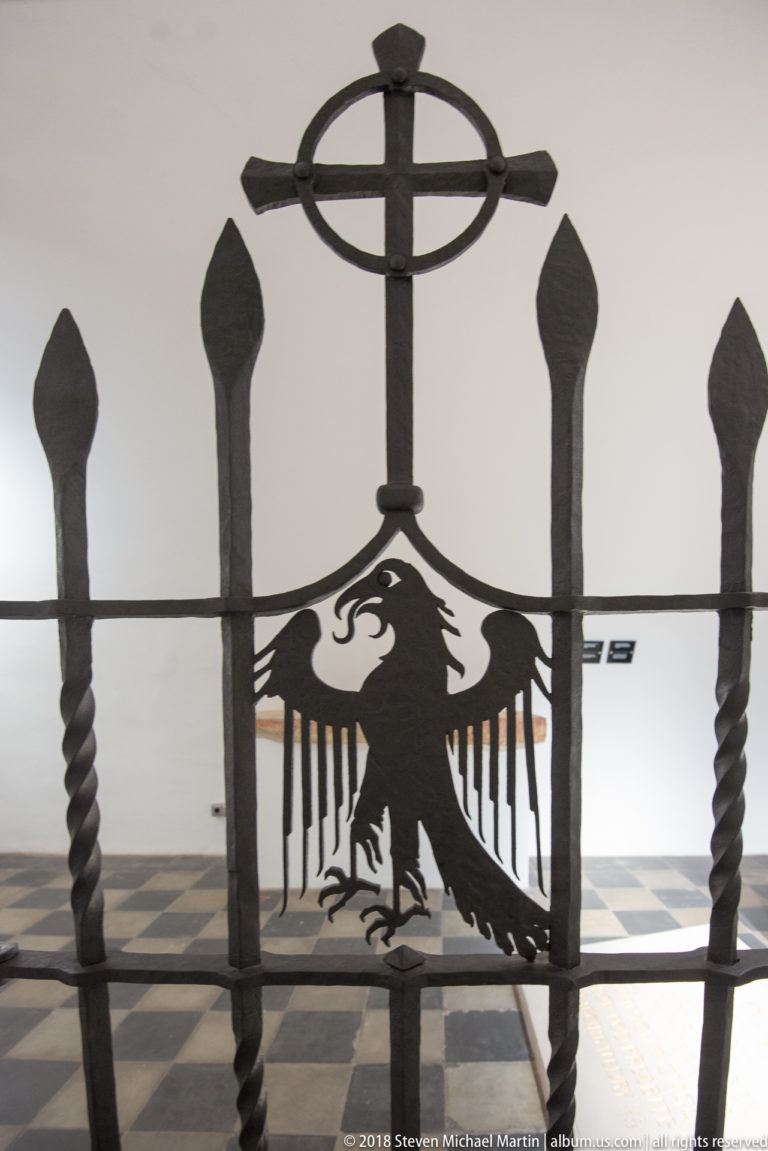 Black wrought-iron gate with cross, stylized Polish eagle, pointed finials, and twisted vertical bars.