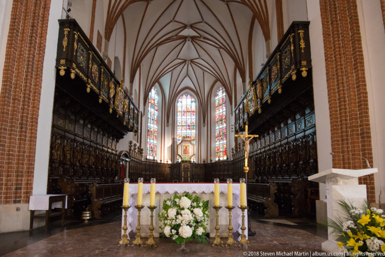 St. John's Archcathedral sanctuary with dark carved choir stalls, stained glass, crucifix, candles, and white flowers.