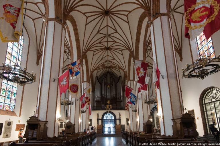 St. John's Archcathedral nave with Gothic rib vaults, brick piers, hanging heraldic banners, pews, and organ loft.
