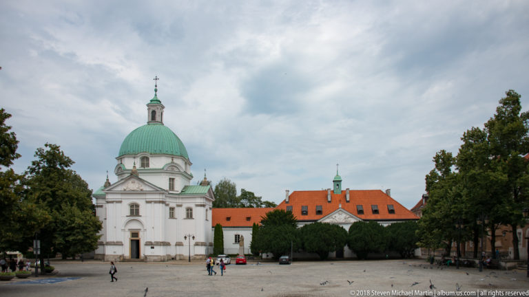 White St. Casimir Church with large green dome and lantern beside red-roofed convent buildings on New Town Market Square.