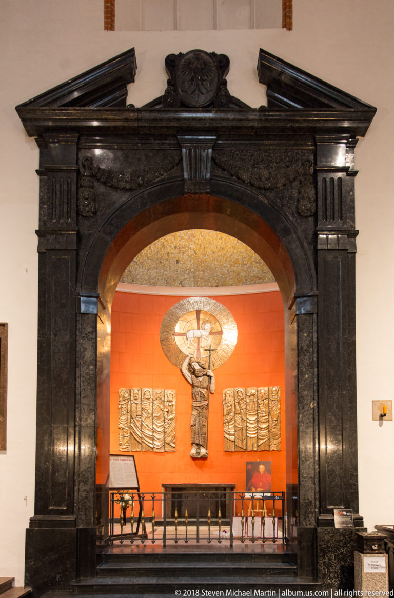 Dark stone arched portal framing a red memorial chapel with relief panels, altar furniture, and a central sculptural cross.