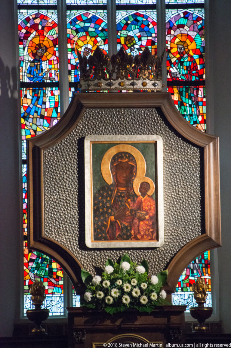 Crowned Black Madonna-style Marian icon in an ornate shrine with white flowers and colorful stained glass behind.