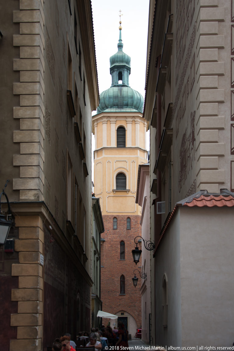 Narrow Warsaw Old Town lane between tall buildings, ending at a pale tower with a green copper roof.