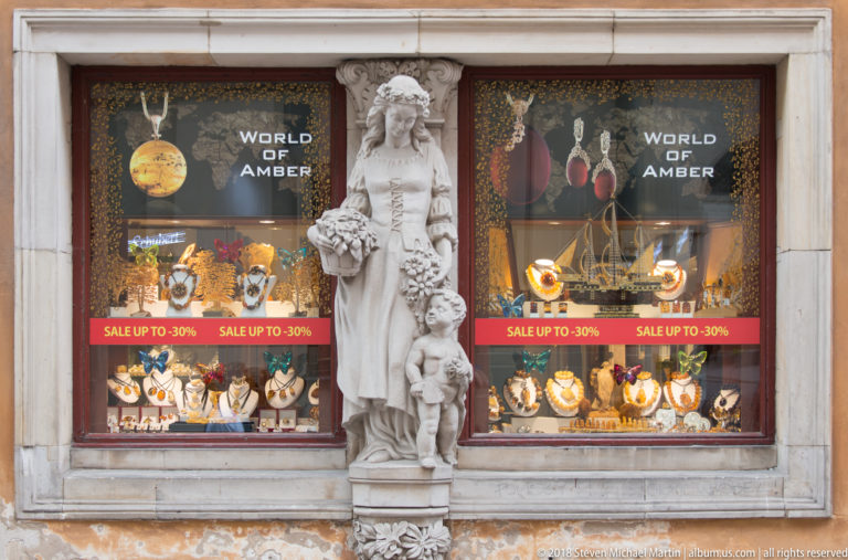 Carved female figure with fruit stands between two shop windows displaying amber jewelry and World of Amber signs.