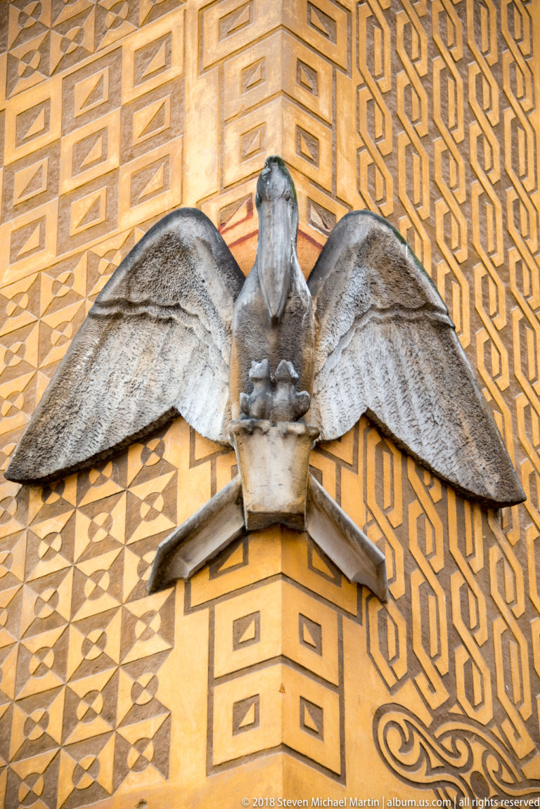Stone eagle with spread wings mounted on an ochre facade covered with geometric decorative patterns.