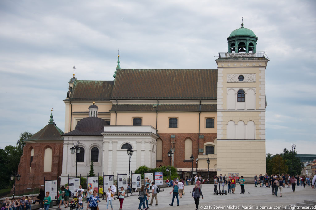 Large Warsaw church with steep roof, pale square bell tower, green cupola, and crowds gathered in the open square.