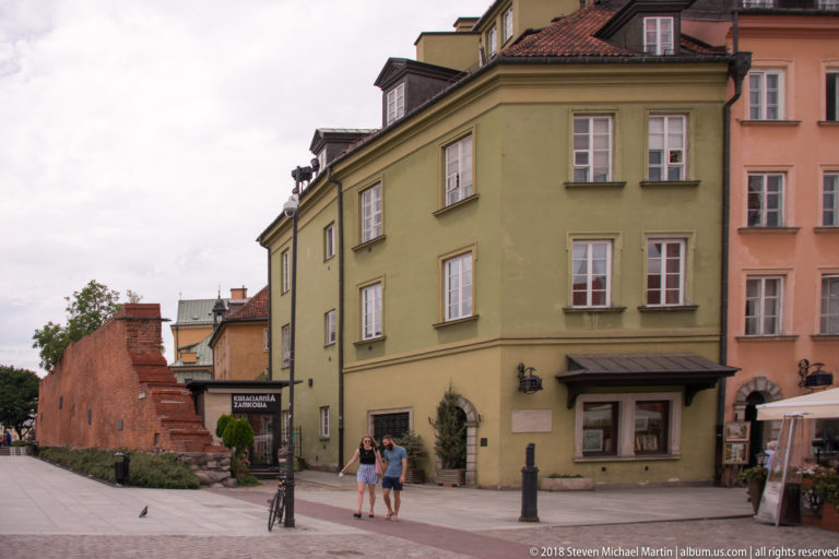 Green historic townhouse beside a red-brick wall and pedestrian lane near Warsaw's Castle Square.