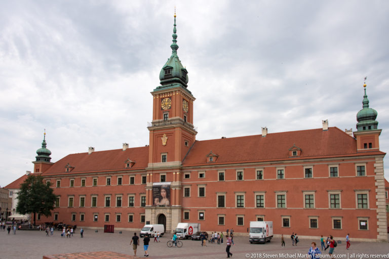 Royal Castle in Warsaw with red facade, central clock tower, green-capped corner towers, and visitors crossing Castle Square.