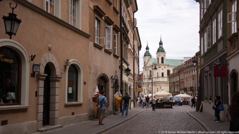 Cobbled Warsaw New Town street lined with historic buildings and pedestrians, ending at a pale twin-towered church.