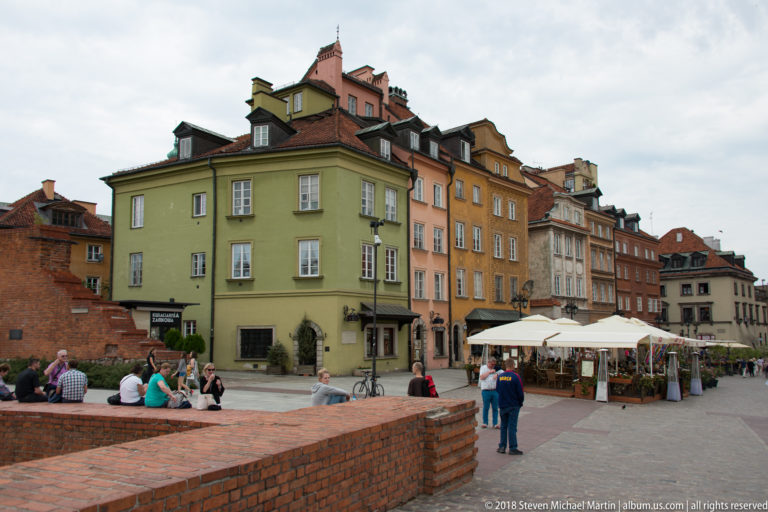Pastel historic townhouses, café umbrellas, pedestrians, and low brick walls beside Warsaw's Castle Square.