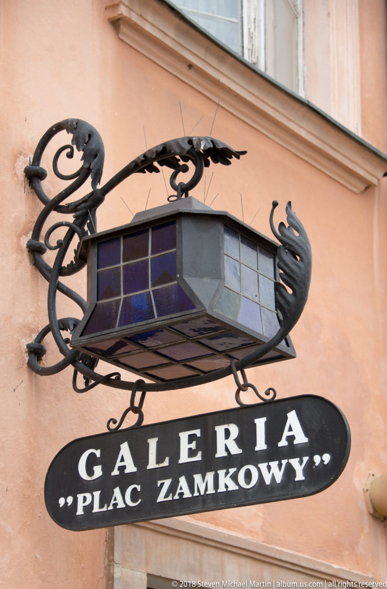 Ornate wrought-iron lantern and oval sign reading Galeria Plac Zamkowy on a peach-colored wall.