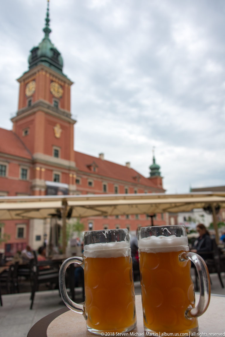 Two mugs of amber beer on an outdoor table with the Royal Castle blurred in the background.