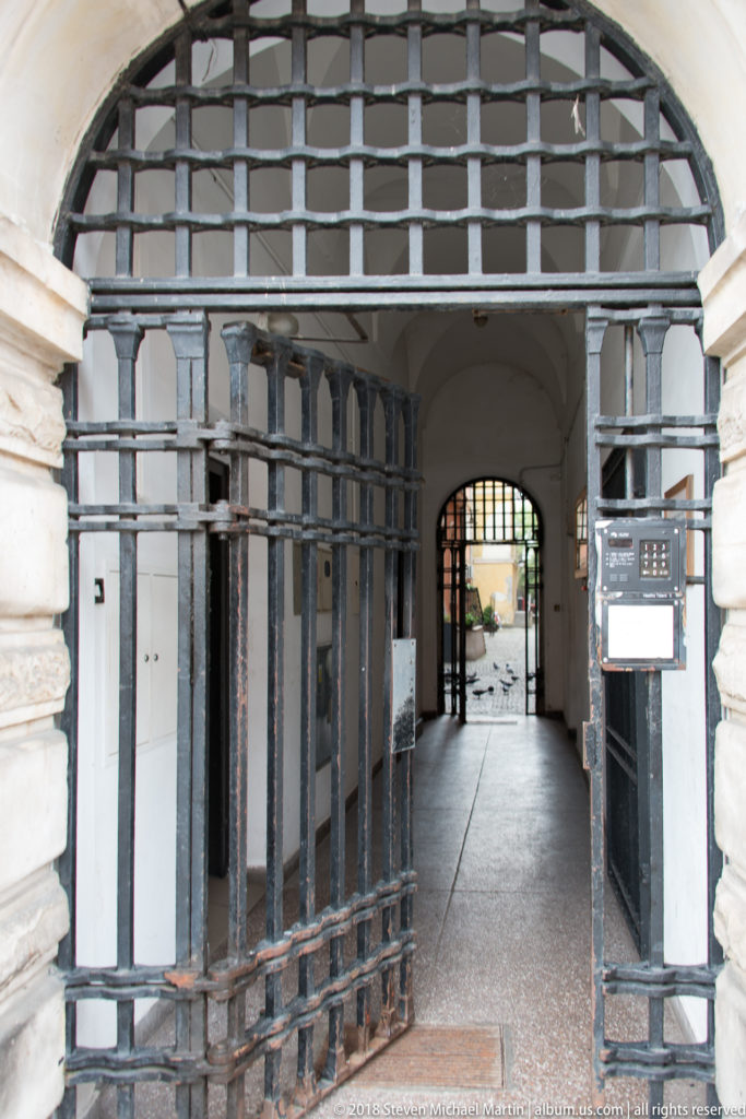 Open iron gate within a rusticated stone archway leading through a covered passage to a bright courtyard.