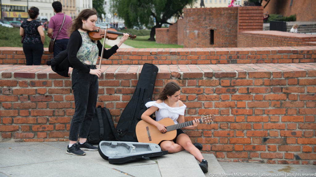 Young violinist and seated guitarist perform beside red-brick Old Town fortifications with open instrument cases.