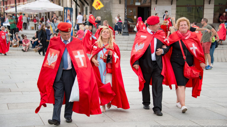 Men and women in bright red ceremonial mantles with white crosses and Polish eagle emblems walk in a Warsaw procession.