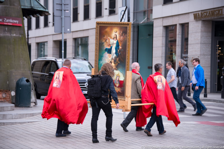 People in red ceremonial cloaks carry a large framed Marian devotional painting through a Warsaw street.