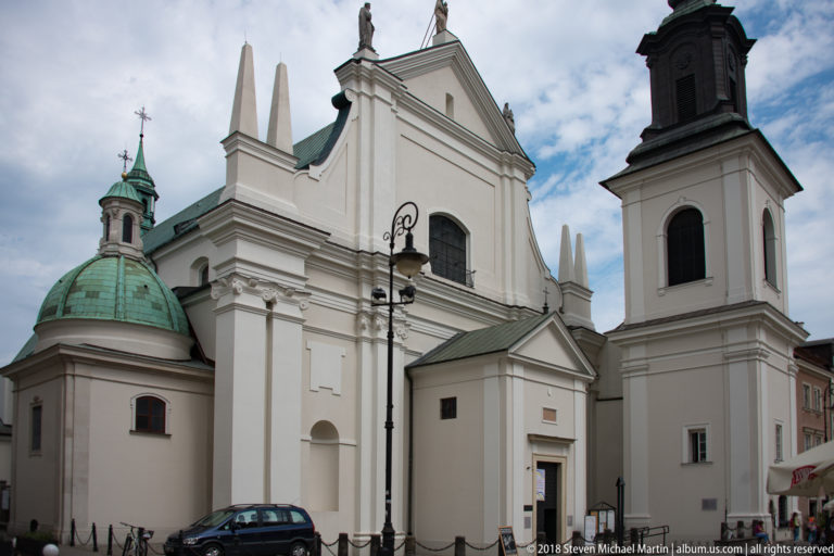 White historic church with central gable, rounded green-domed chapel, and square bell tower under a cloudy sky.