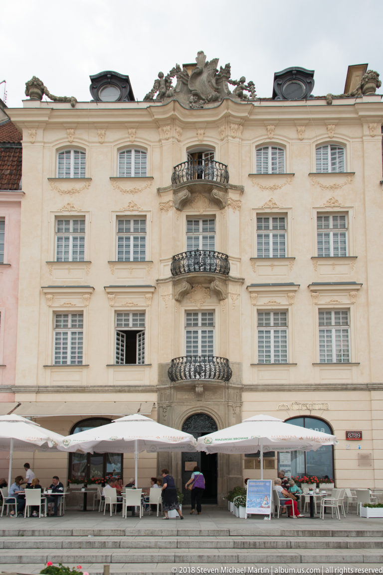 Ornate pale townhouse with curved balconies, sculpted roofline, tall windows, and outdoor café tables at street level.
