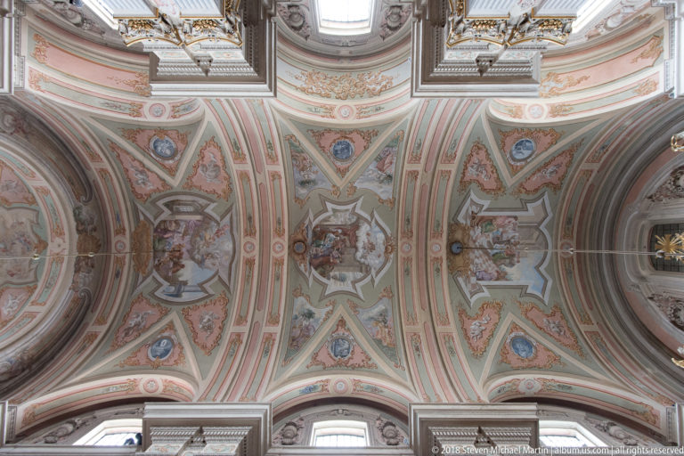 Pastel-painted Baroque church ceiling with figurative frescoes, medallions, and ornate architectural borders.