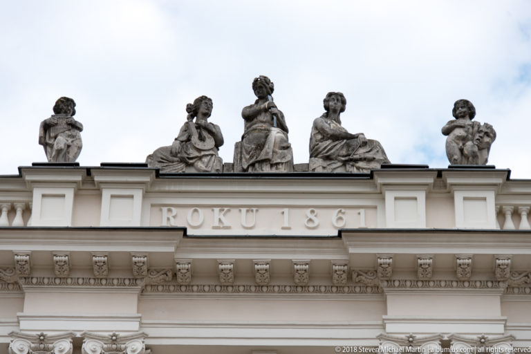 Five stone female statues stand on a roofline above a balustrade and the carved inscription ROKU 1861.
