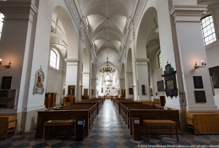 Long white church nave with checkered floor, wooden pews, wall monuments, and a bright sanctuary in the distance.