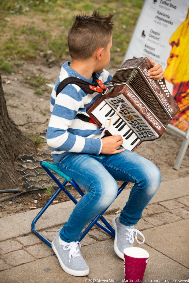 Young street musician seated on a folding chair playing a large accordion beside a donation cup.