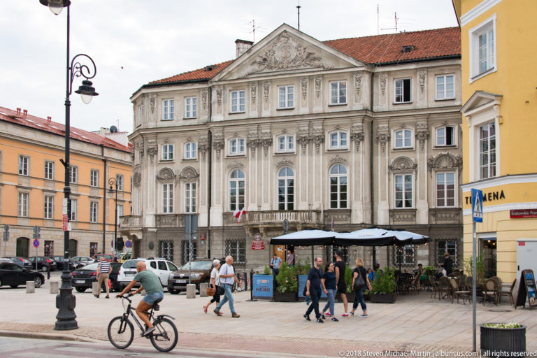 Ornate cream historic building with pediment and tall windows behind cyclists, pedestrians, restaurant seating, and parked cars.
