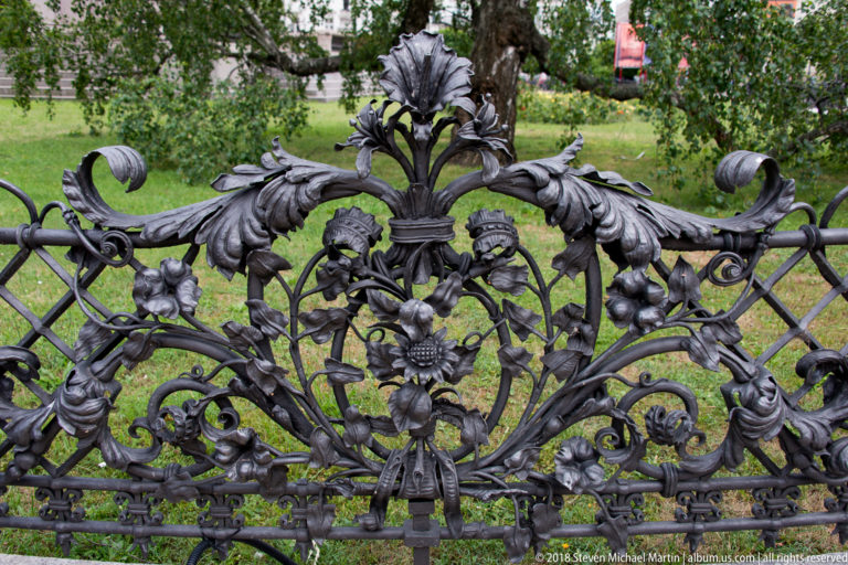 Close view of ornate wrought-iron fence with leaves, flowers, scrolls, and crest-like motifs around the Mickiewicz Monument.