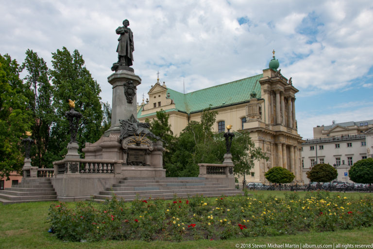 Adam Mickiewicz Monument with standing bronze figure, flower beds, balustrade, and the Carmelite Church behind.