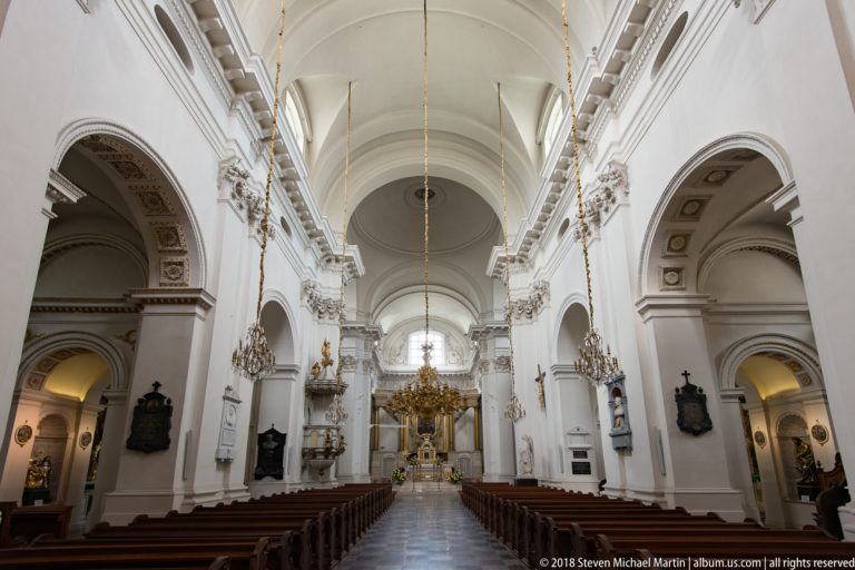 Long white Carmelite Church nave with wooden pews, side chapels, chandeliers, and the high altar in the distance.