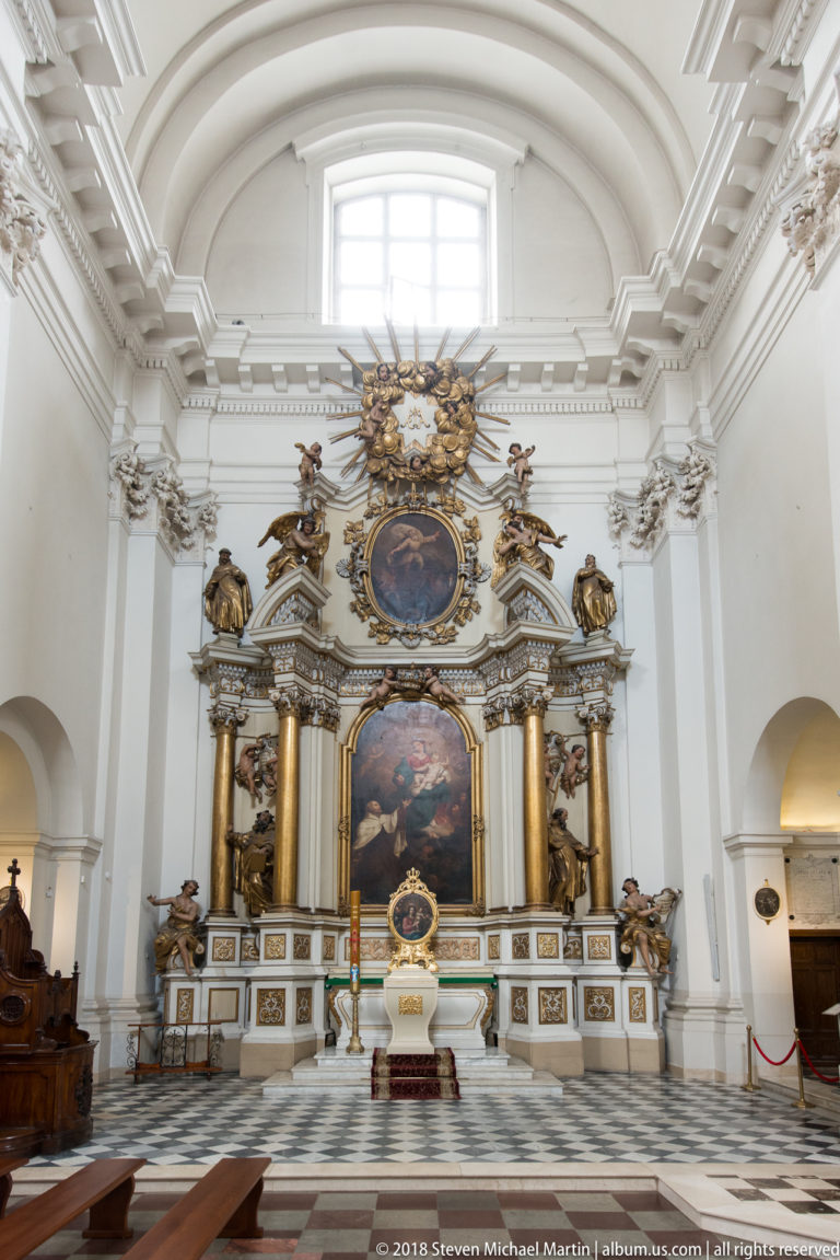 Baroque side altar with large paintings, gilded columns, sculpted figures, and a black-and-white checkered floor.