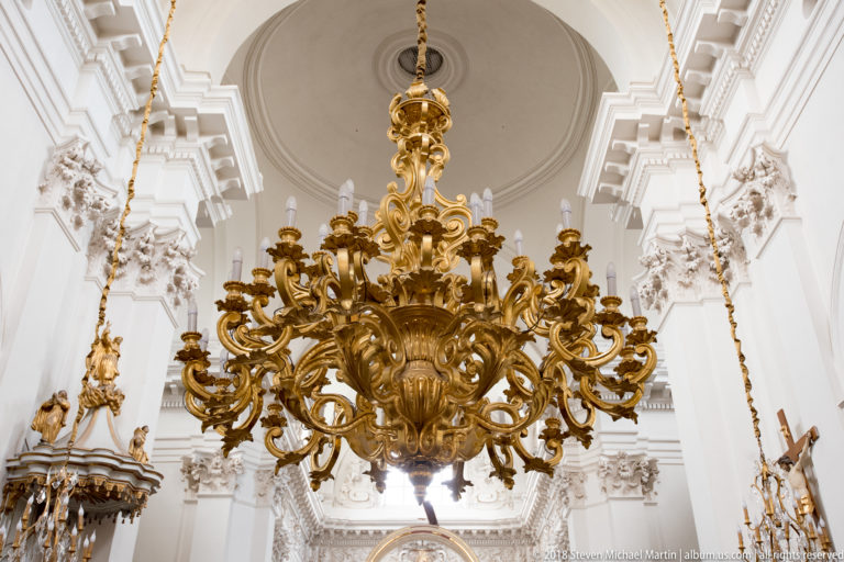 Large gilded Rococo chandelier with many scrolling candle arms suspended beneath a white vaulted church ceiling.