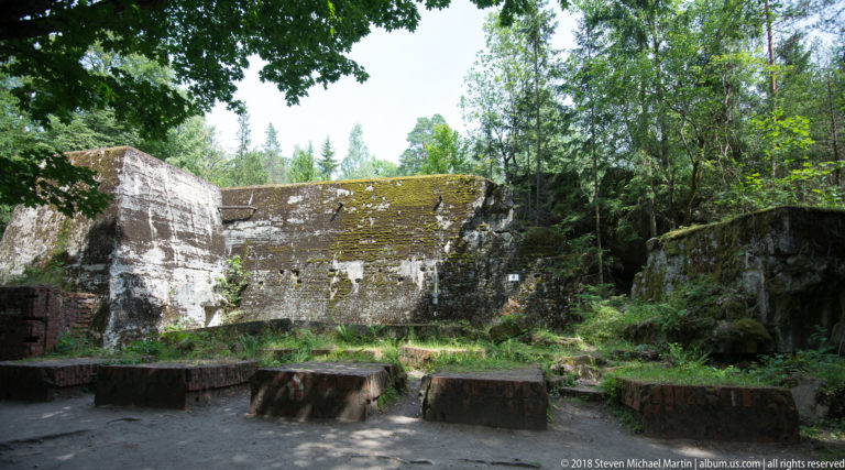 Open ruin enclosed by massive mossy concrete walls, with low rectangular concrete blocks arranged in the foreground.
