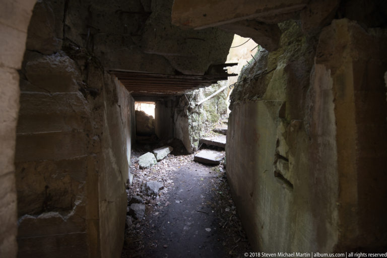 Dark concrete corridor filled with rubble and collapsed slabs, with daylight entering through broken openings.