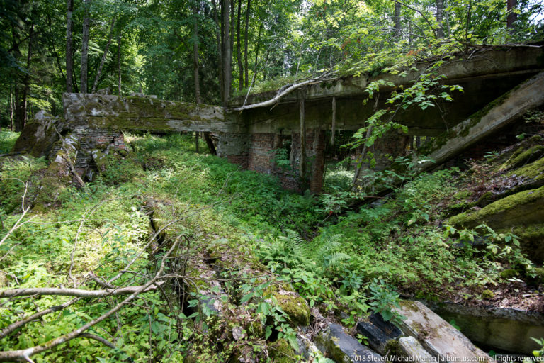 Long broken concrete structure on supports crossing an overgrown depression, with one end collapsed toward the forest floor.