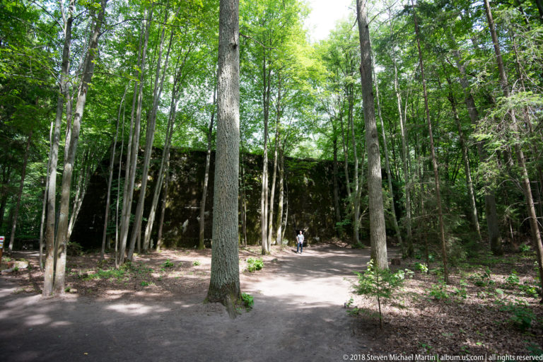 Tall trees screen a massive dark concrete bunker wall beside a forest path, with a small visitor providing scale.