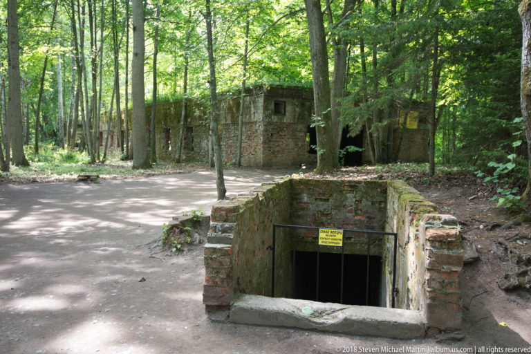 Brick-lined stair opening descending below ground, with a low ruined building and trees behind it.