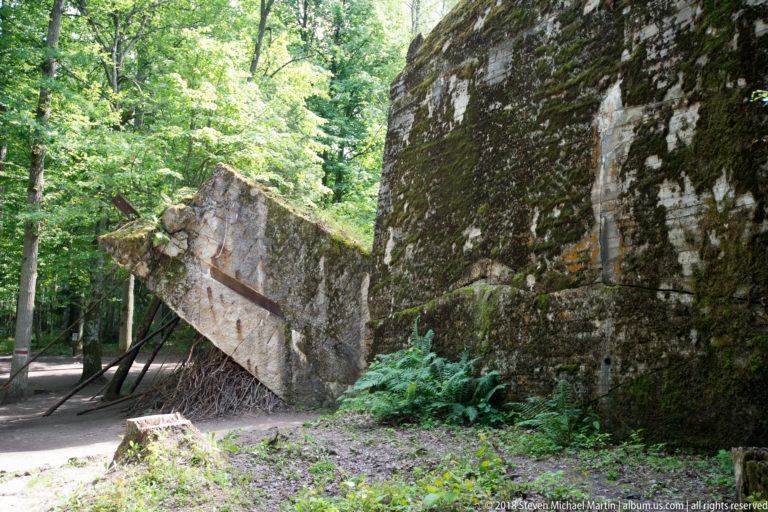 Huge broken concrete slab leaning diagonally beside a standing bunker wall, supported from below in dense woodland.