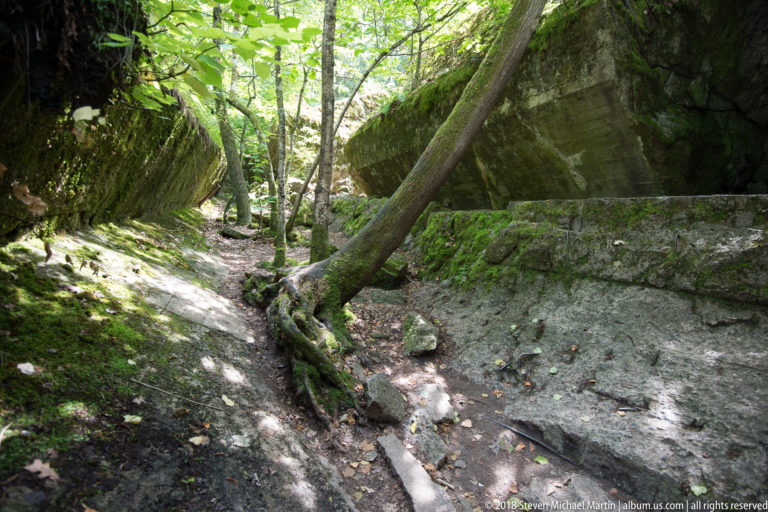 Large mossy concrete slab tilted over a narrow depression, with trees and broken masonry beneath and around it.