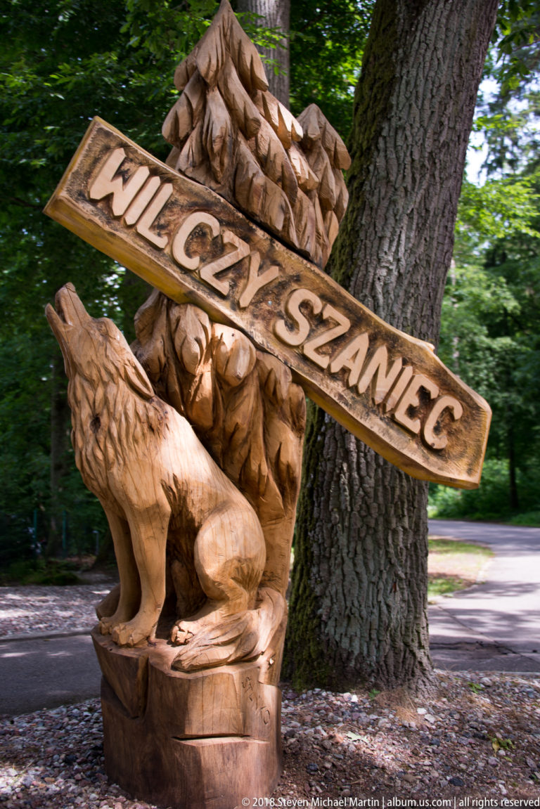 Carved wooden WILCZY SZANIEC sign above a sculpted wolf beside a large tree in the forested visitor area.