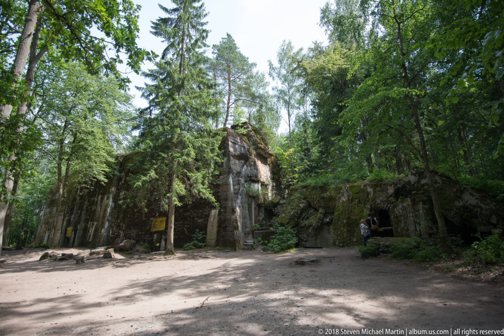 Wide forest clearing with large bunker ruins, dark openings, broken concrete, and visitors near structure 13.